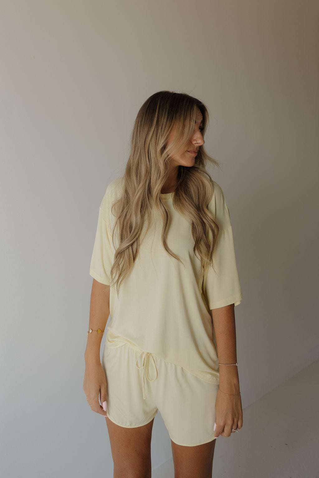 A woman with long, wavy blonde hair stands against a plain light-colored background, looking to her left, wearing the Women’s Bamboo Short Set in Sunny by forever french baby.