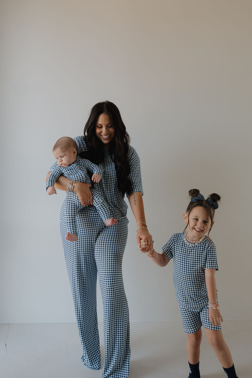 A woman with a baby and young girl, all in matching forever french baby Bamboo Short Set | Blue Gingham pajamas, stand against a light background—perfect for showcasing breathable bamboo family sleepwear.