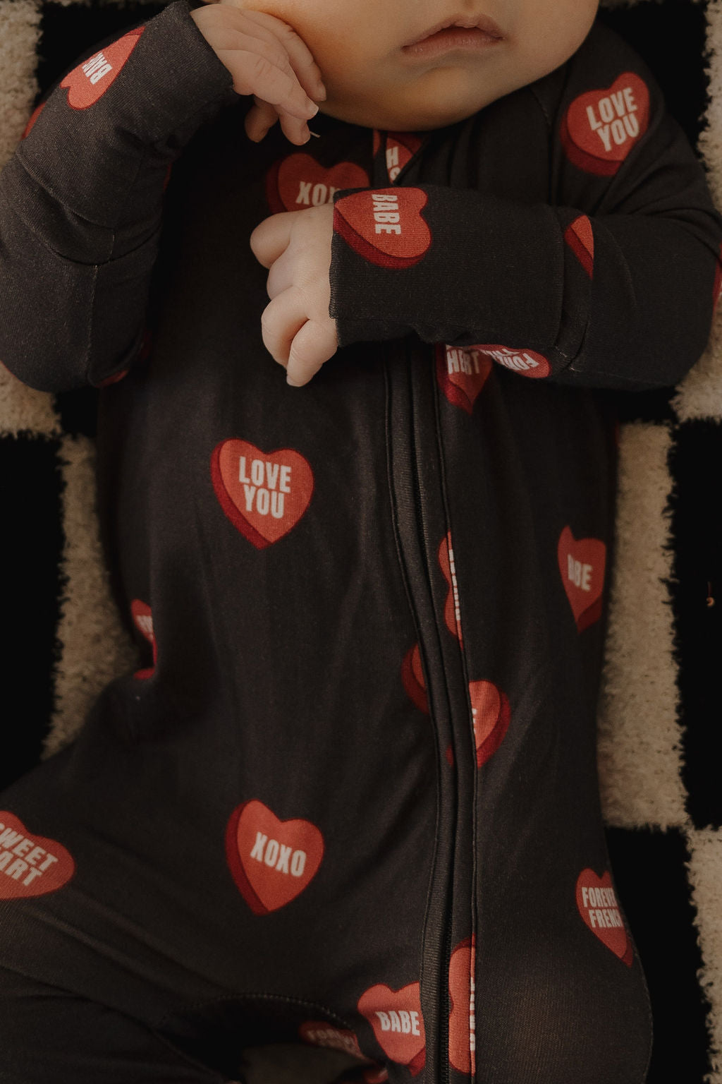A baby wears forever french baby's Bamboo Zip Pajamas | Love Notes – Charcoal, a hypo-allergenic black onesie with red heart-shaped “LOVE YOU,” “XOXO,” and “Babe” messages. The baby's face is partly visible with one hand near their mouth.