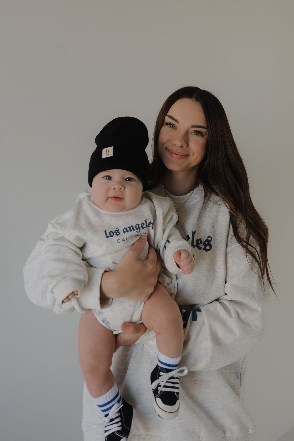 A woman in a light gray sweatshirt holds a smiling baby wearing the forever french baby Sweatshirt Romper by ff Los Angeles, paired with a black beanie and sneakers. They pose against a plain light gray background.