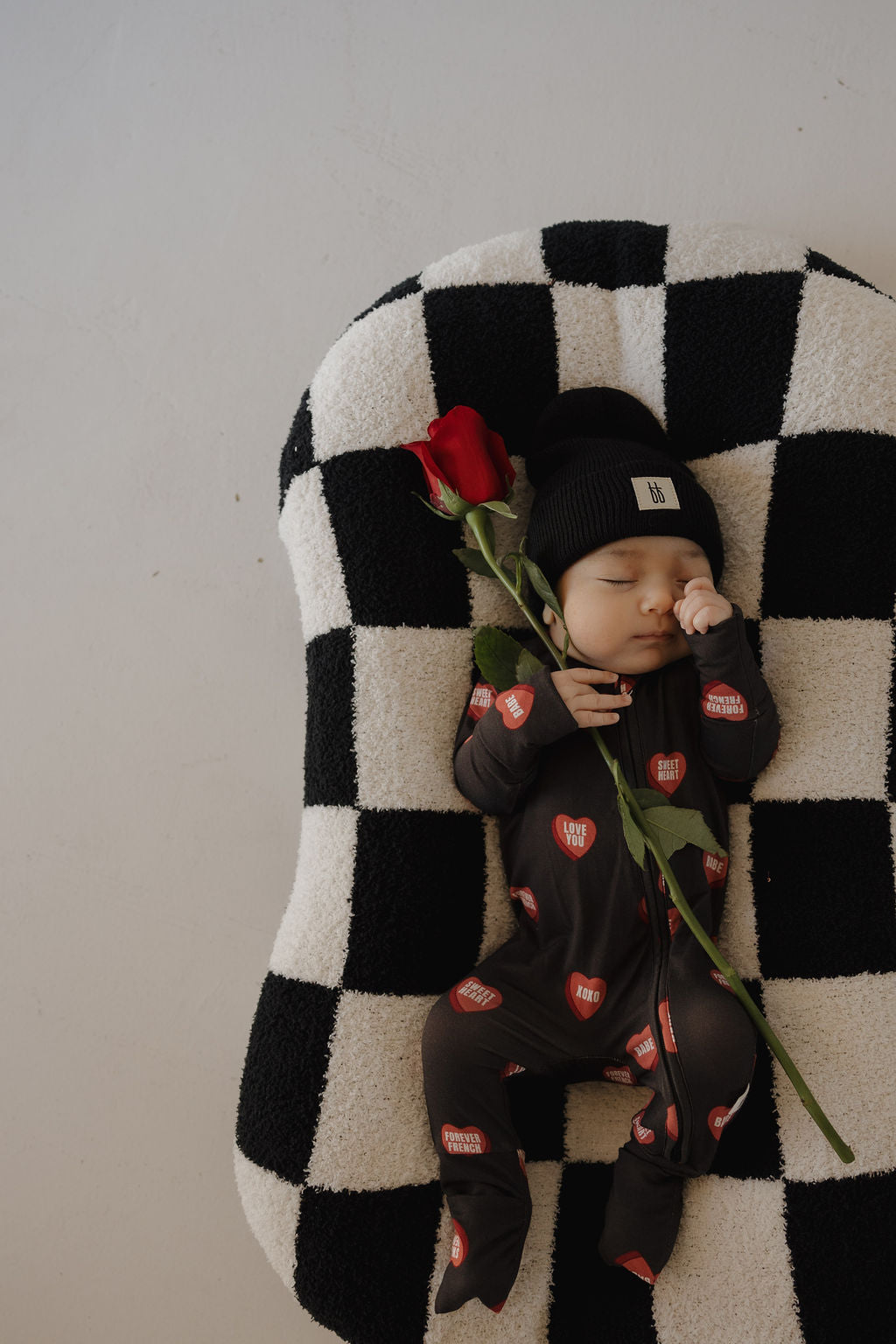 A baby in forever french baby’s Bamboo Zip Pajamas | Love Notes – Charcoal and a black beanie sleeps on a black-and-white checkered cushion, holding a single red rose.