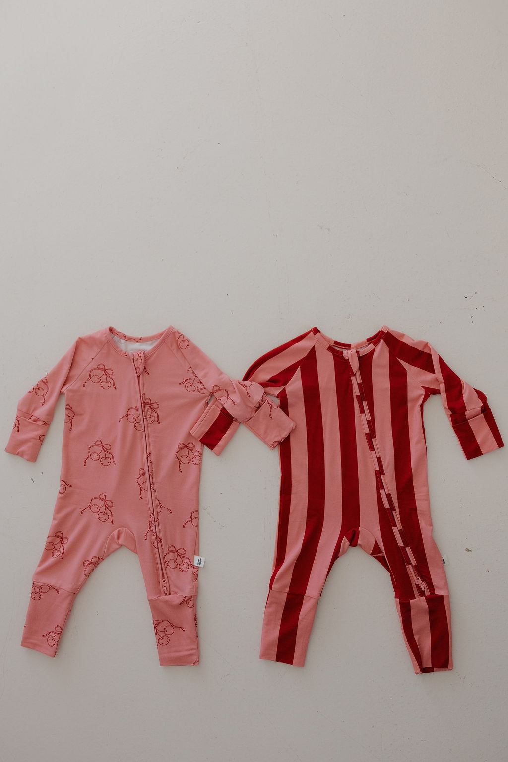 Two Minty Cherries Bamboo Zip Pajamas laid flat: one pink with red berries, and one red with wide pink stripes. Both feature front zippers and are made from breathable bamboo; displayed side by side on a light background.