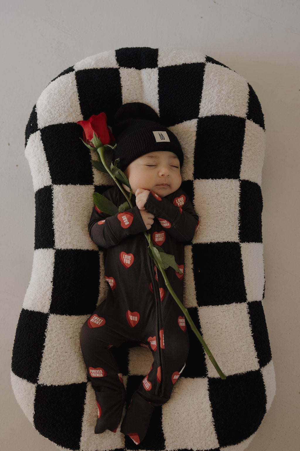 A baby in forever french baby’s Bamboo Zip Pajamas | Love Notes – Charcoal and a black beanie sleeps on a black-and-white checkered cushion, holding a single red rose.