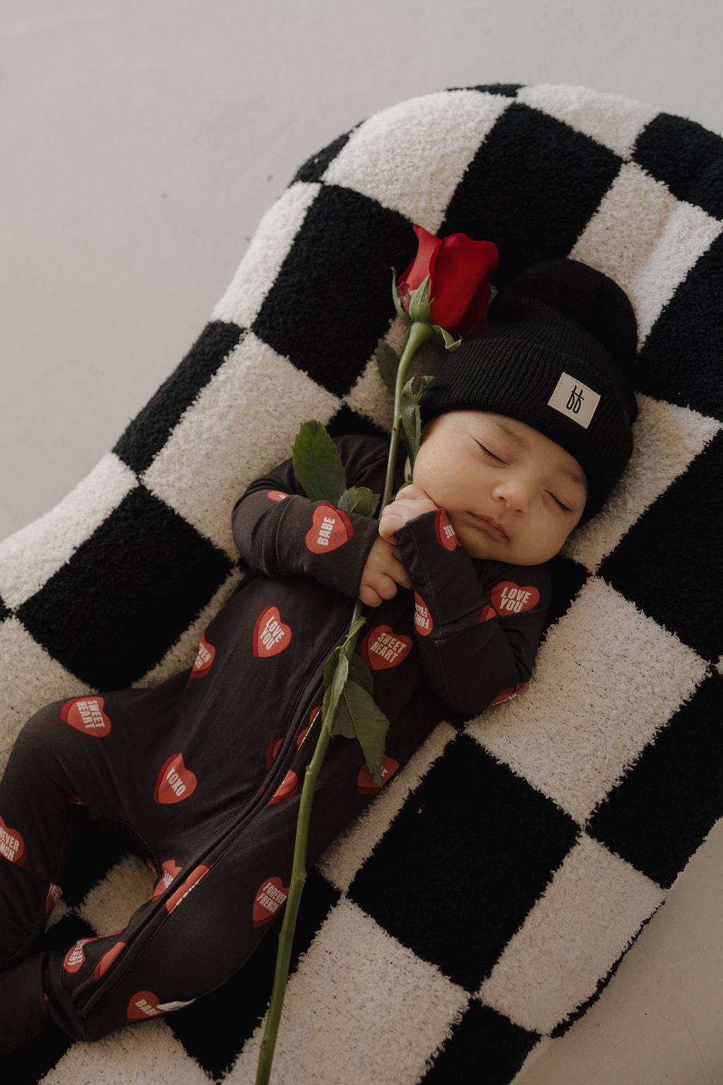 A baby in forever french baby's Bamboo Zip Pajamas | Love Notes – Charcoal sleeps on a black-and-white checkered blanket, wearing a black beanie and holding a single red rose.