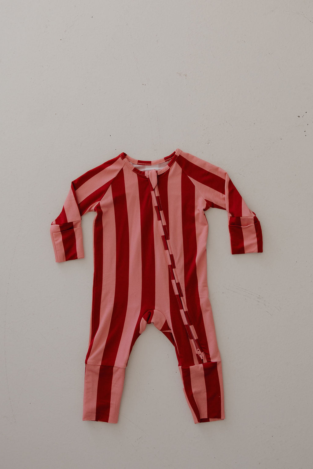 The Minty Cherries Bamboo Zip Pajamas | Iconic Stripe is a hypo-allergenic baby romper with long sleeves and legs, featuring bold red and pink vertical stripes, a diagonal zipper, and breathable fabric for comfortable sleep. Laid flat on a light background.