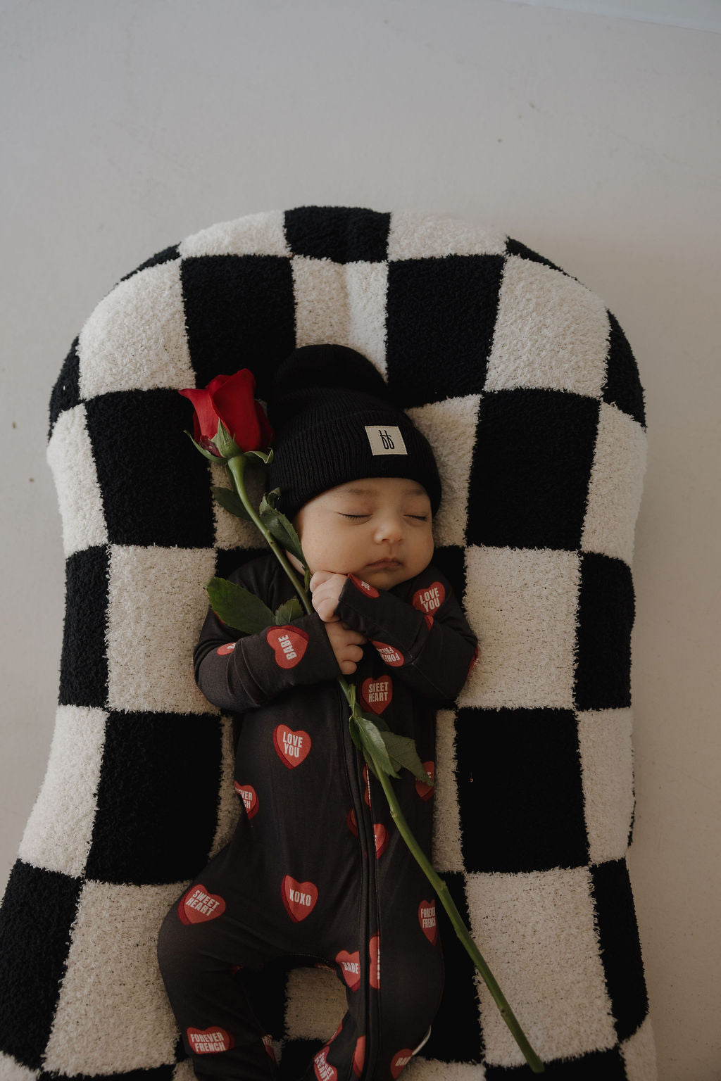A baby sleeps peacefully in forever french baby’s Bamboo Zip Pajamas | Love Notes – Charcoal, with red heart patches and a black beanie, resting on a checkered cushion and holding a single red rose.