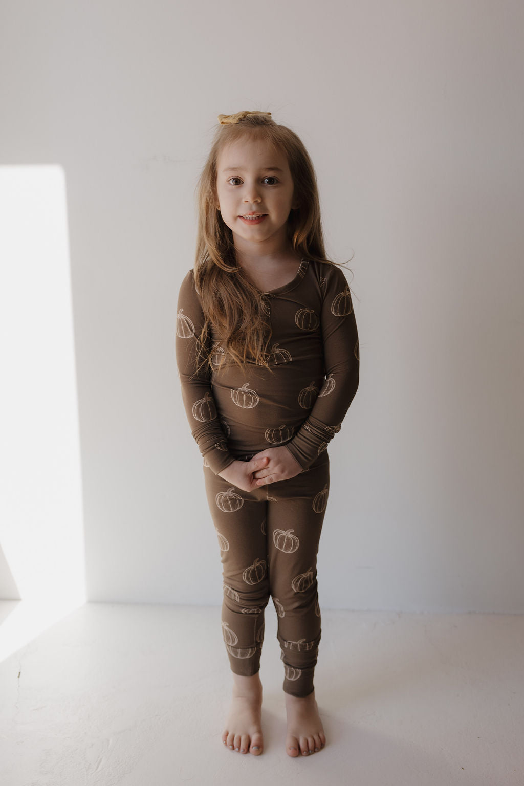 A young girl with long hair, barefoot and smiling, stands against a light wall wearing forever french baby’s Bamboo Two Piece Pajamas | Brown Pumpkin—hypoallergenic kids pajamas designed for comfort and sensitive skin.