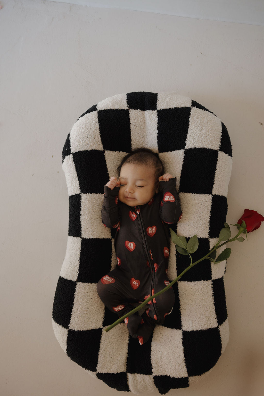 A baby in forever french baby's Bamboo Zip Pajamas | Love Notes – Charcoal, featuring red heart patterns, sleeps on a black-and-white checkered cushion while holding a single red rose.