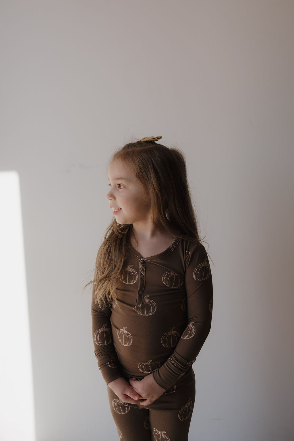 A young girl with long hair stands against a plain light wall, looking to the side. She wears forever french baby Bamboo Two Piece Pajamas | Brown Pumpkin and has her hands clasped in front of her.