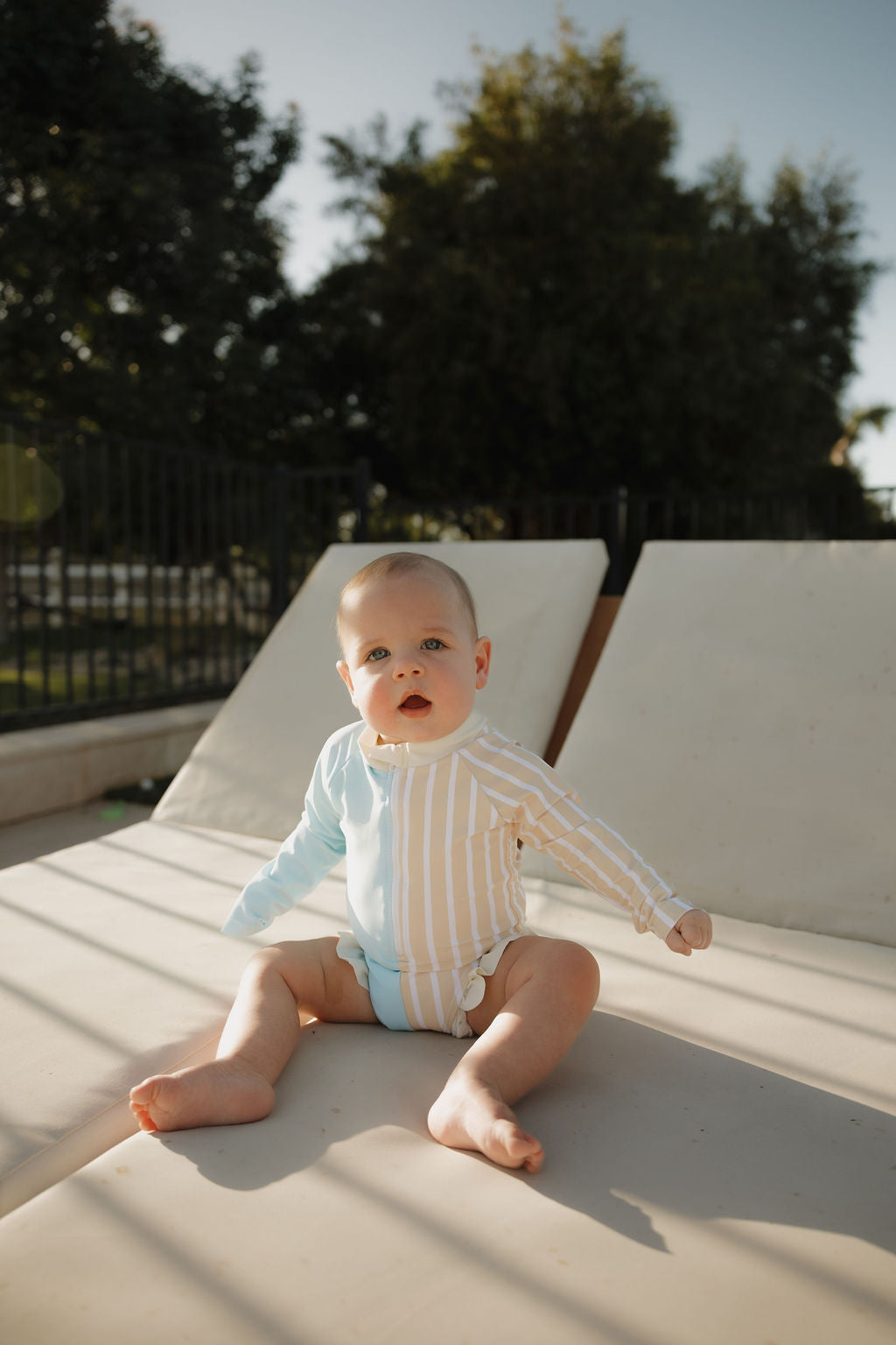 A baby in a lolo webb 2 Girl Long Sleeve Swimsuit | Surf & Sand sits on a lounge chair outdoors, with trees and sunlight in the background.