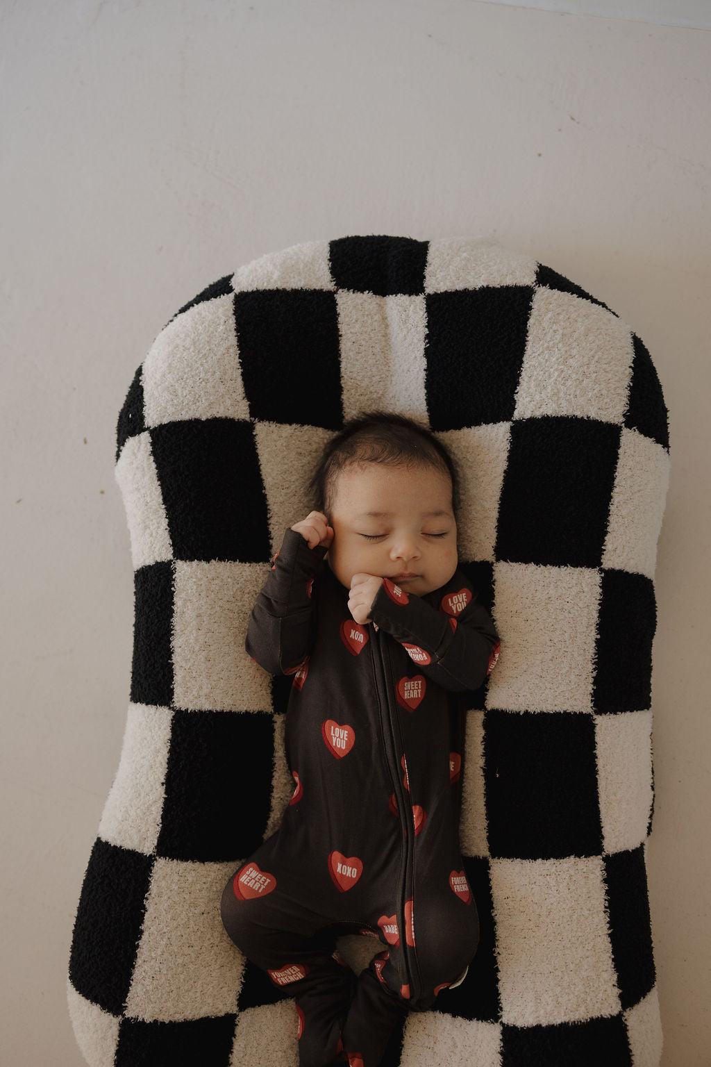 A baby sleeps peacefully on a black and white checkered cushion, wearing forever french baby Bamboo Zip Pajamas | Love Notes – Charcoal, a hypo-allergenic onesie with red hearts and white lettering.