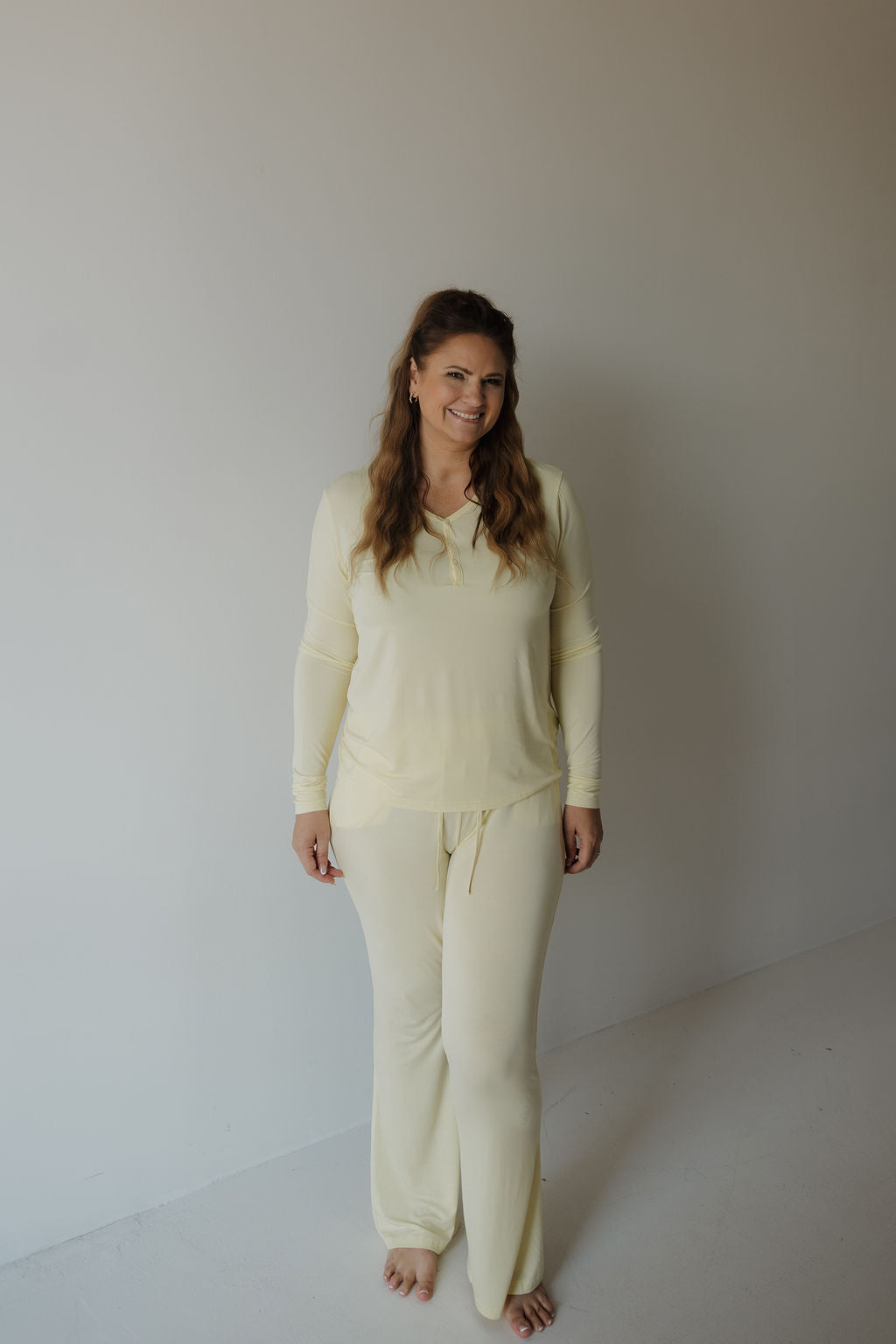 A woman with light brown, loosely waved hair stands barefoot indoors against a plain white wall and floor, smiling as she wears the forever french baby Women’s Long Sleeve Bamboo Pajama in Sunny—a light yellow, hypo-allergenic set featuring long sleeves and matching pants designed for ultimate comfort.