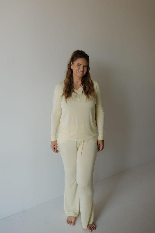 A woman with light brown, loosely waved hair stands barefoot indoors against a plain white wall and floor, smiling as she wears the forever french baby Women’s Long Sleeve Bamboo Pajama in Sunny—a light yellow, hypo-allergenic set featuring long sleeves and matching pants designed for ultimate comfort.