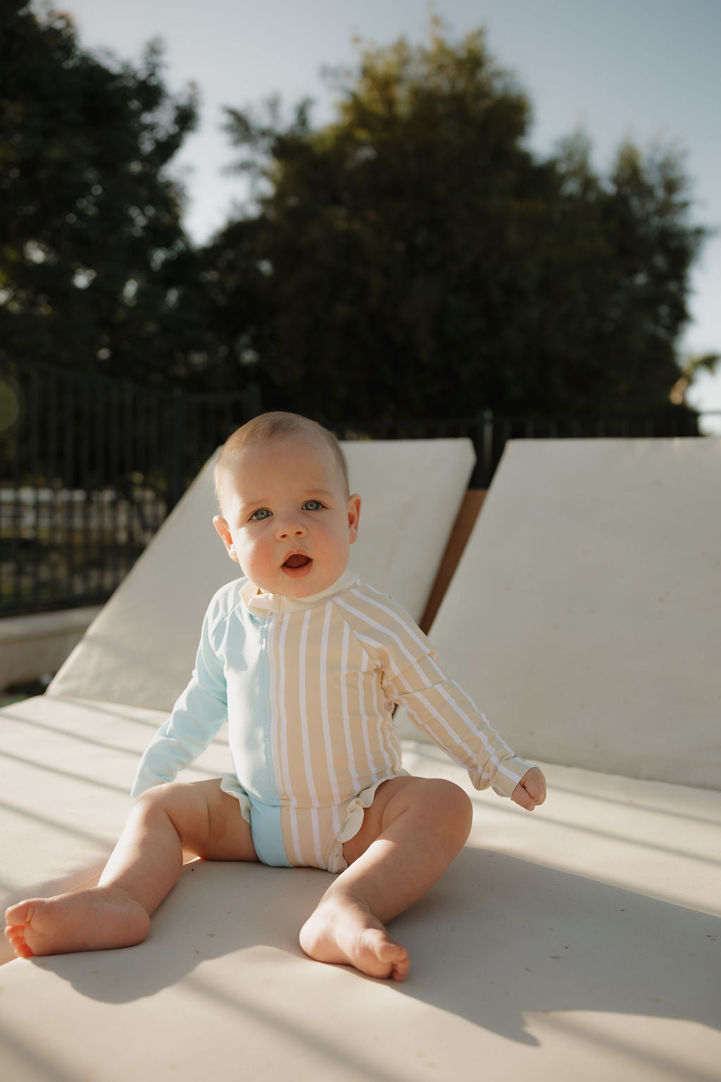 A baby wearing the lolo webb 2 Girl Long Sleeve Swimsuit | Surf & Sand sits on a lounge chair outdoors, gazing curiously to the side as sunlight filters through the trees in the background.