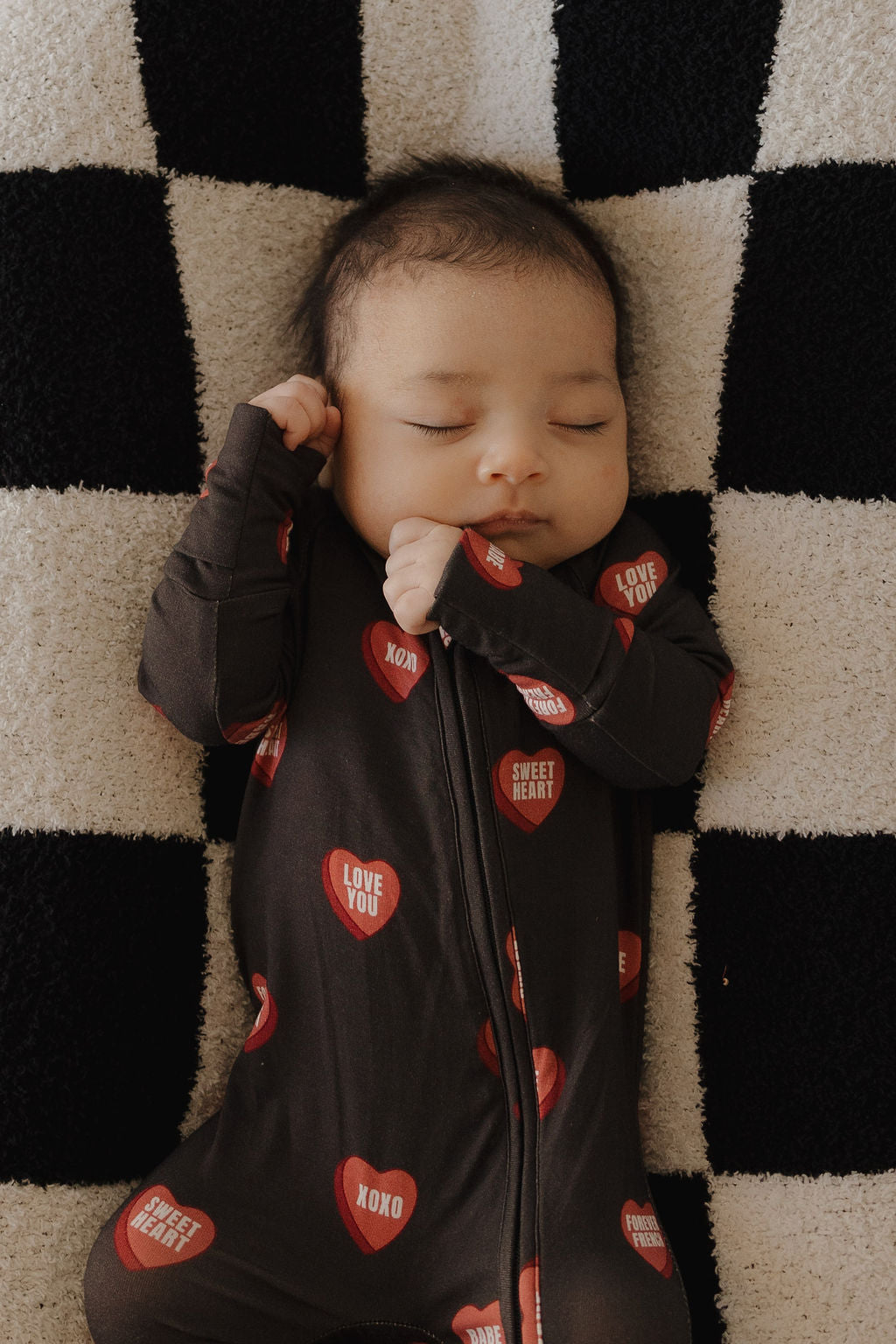 A baby sleeps on a black and white checkered blanket, cozy in forever french baby’s Bamboo Zip Pajamas | Love Notes – Charcoal, a breathable dark onesie with red heart patches that say “Love You,” “XOXO,” and “Sweet Heart.”.