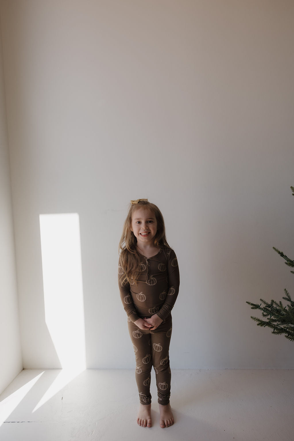A young girl with long hair smiles barefoot against a white wall, wearing forever french baby’s Bamboo Two Piece Pajamas in Brown Pumpkin. Sunlight streams in from the left, with a green plant partly visible on the right.