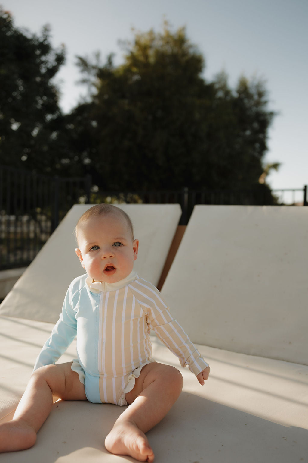 A baby wearing the lolo webb 2 Girl Long Sleeve Swimsuit | Surf & Sand sits outdoors on a lounge chair, surrounded by trees and a fence, enjoying gentle natural sunlight.