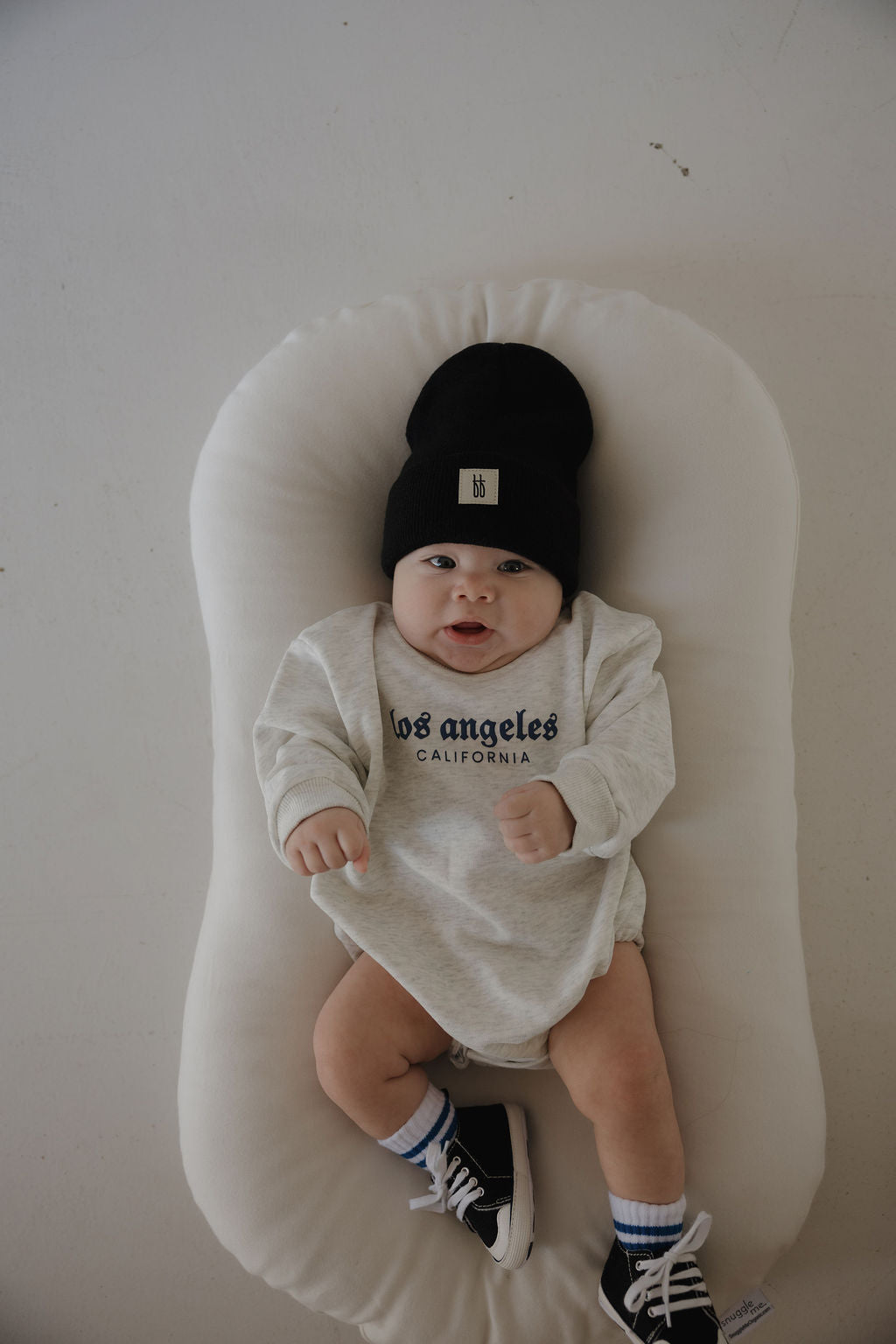 A baby lies on a white cushion, wearing a black beanie, striped socks, black sneakers, and a light gray forever french baby "Sweatshirt Romper | ff Los Angeles," looking up with an open mouth and clenched fists.