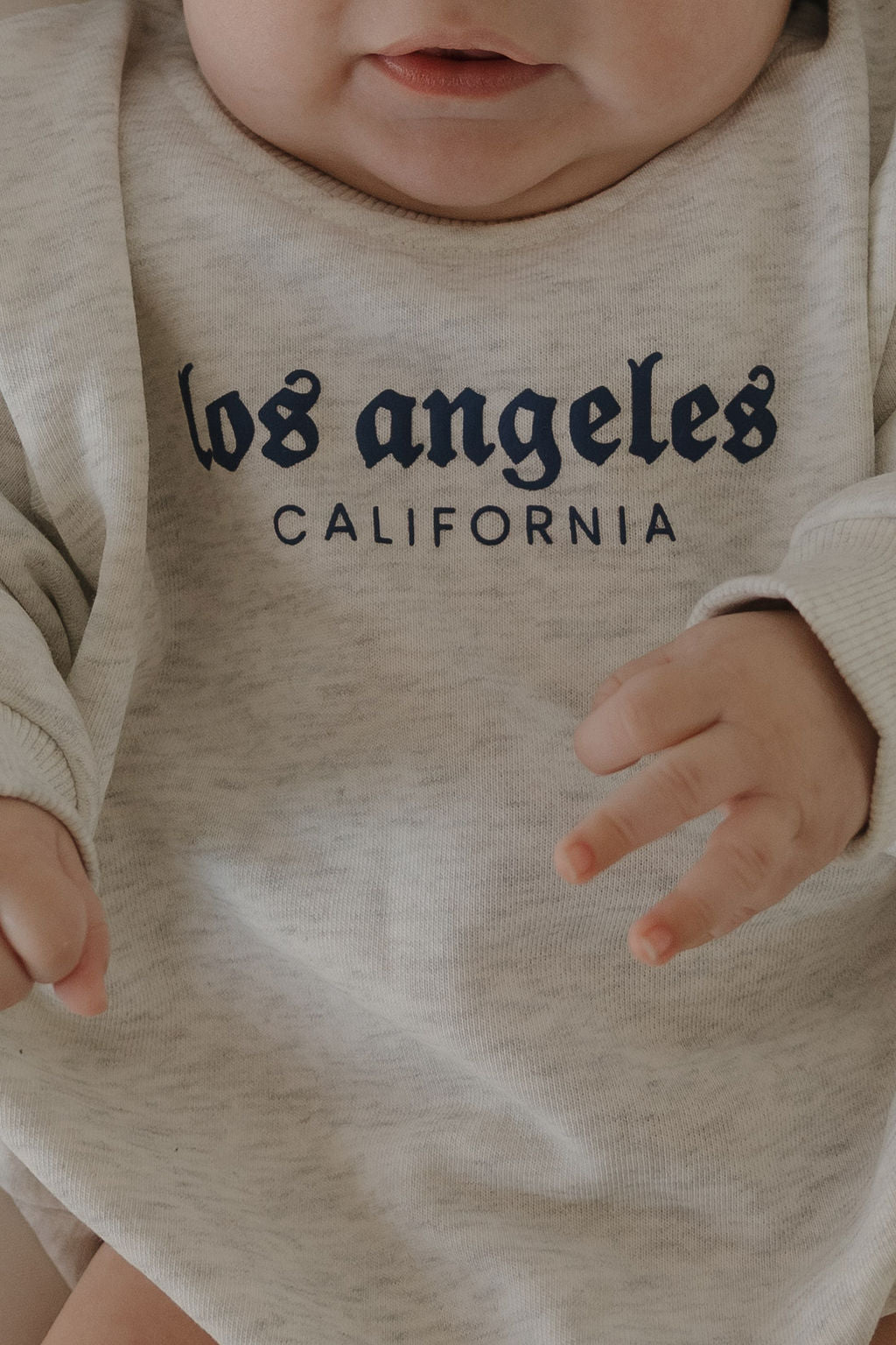 A close-up of a baby wearing the forever french baby Sweatshirt Romper | ff Los Angeles, a light gray romper featuring "los angeles CALIFORNIA" in navy blue on the front. The baby's hands and part of their mouth are visible.