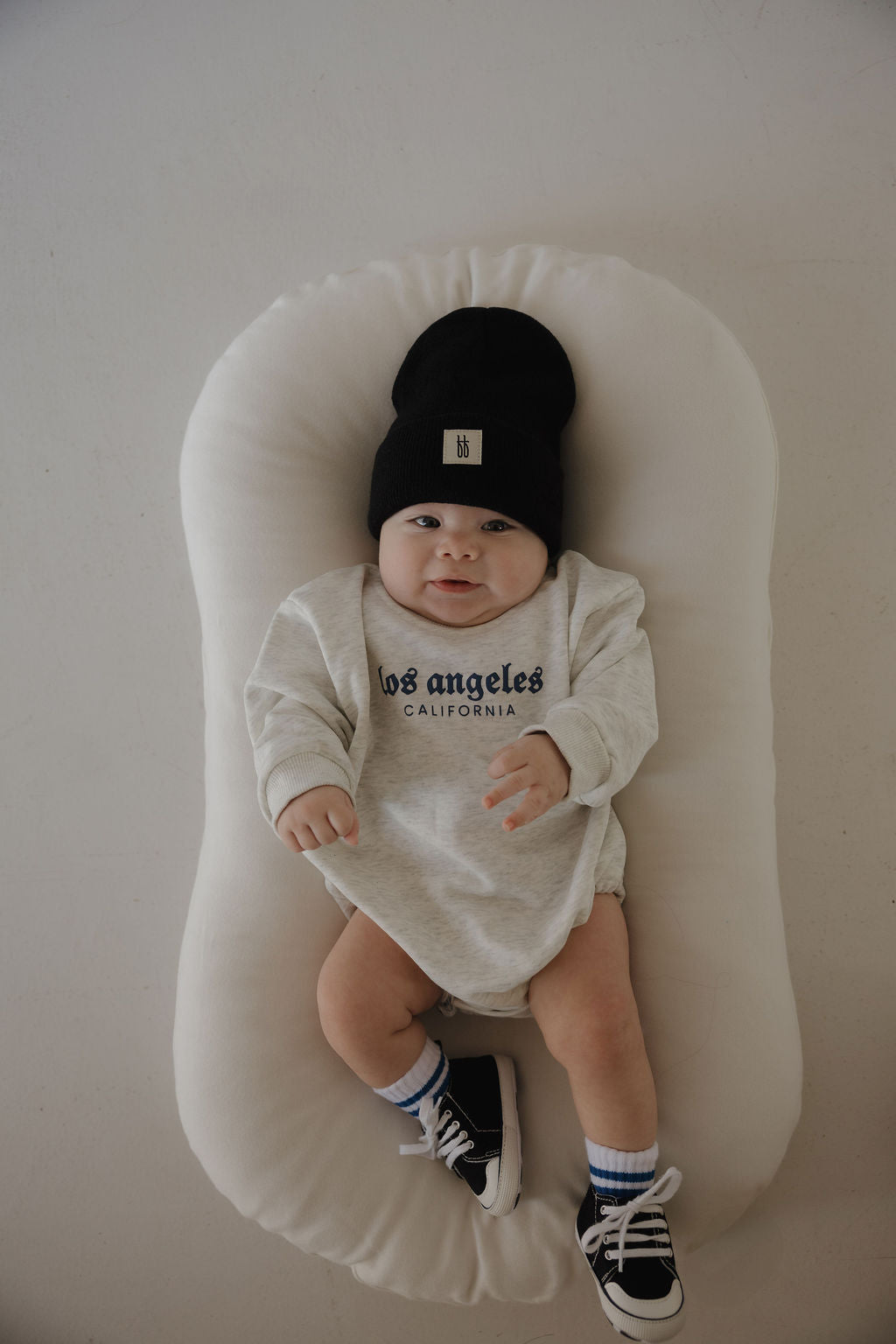 A baby lies on a white cushion wearing a black beanie, striped socks, black sneakers, and the forever french baby Sweatshirt Romper | ff Los Angeles, looking up with one hand raised.
