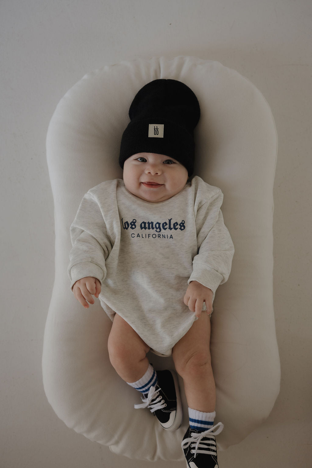 A smiling baby in a black beanie, striped socks, black sneakers, and the forever french baby Sweatshirt Romper | ff Los Angeles lies on a soft white cushion.