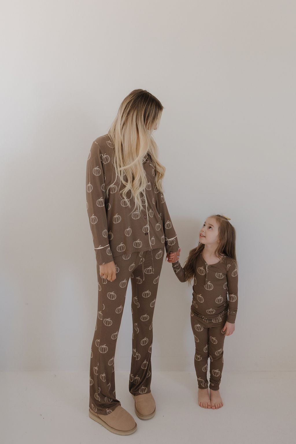 A woman and a young girl wear matching forever french baby Bamboo Two Piece Pajamas in Brown Pumpkin, holding hands and smiling against a white wall—cozy, breathable bamboo sleepwear perfect for special moments together.