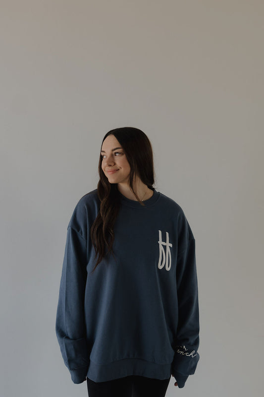 A woman with long dark hair wears a relaxed fit Adult Sweatshirt | ff Blue by forever french baby, featuring a white chest design, as she stands against a light background, looking left and smiling softly.