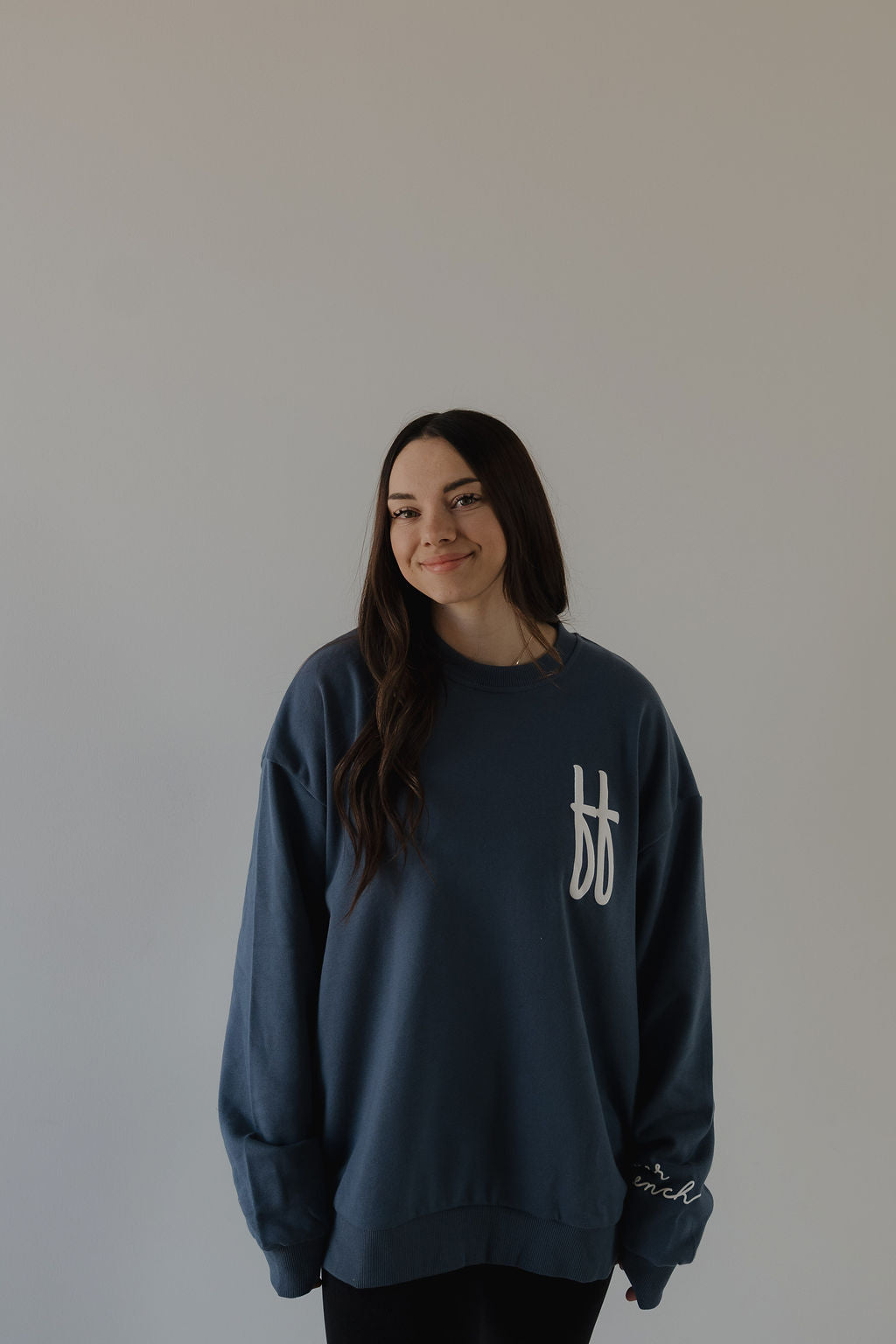 A young woman with long dark hair smiles softly against a plain light wall, wearing a relaxed fit Adult Sweatshirt | ff Blue by forever french baby, featuring a white logo on the chest and sleeve.