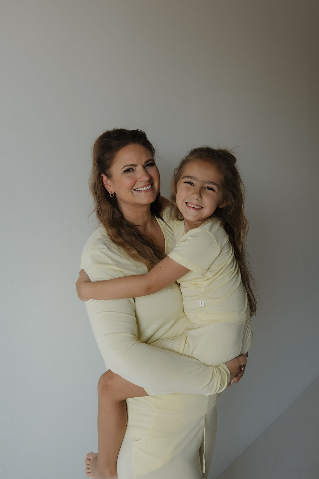 A smiling woman hugs a young girl, both dressed in matching forever french baby Bamboo Short Set | Sunny pajamas, standing against a plain white wall.