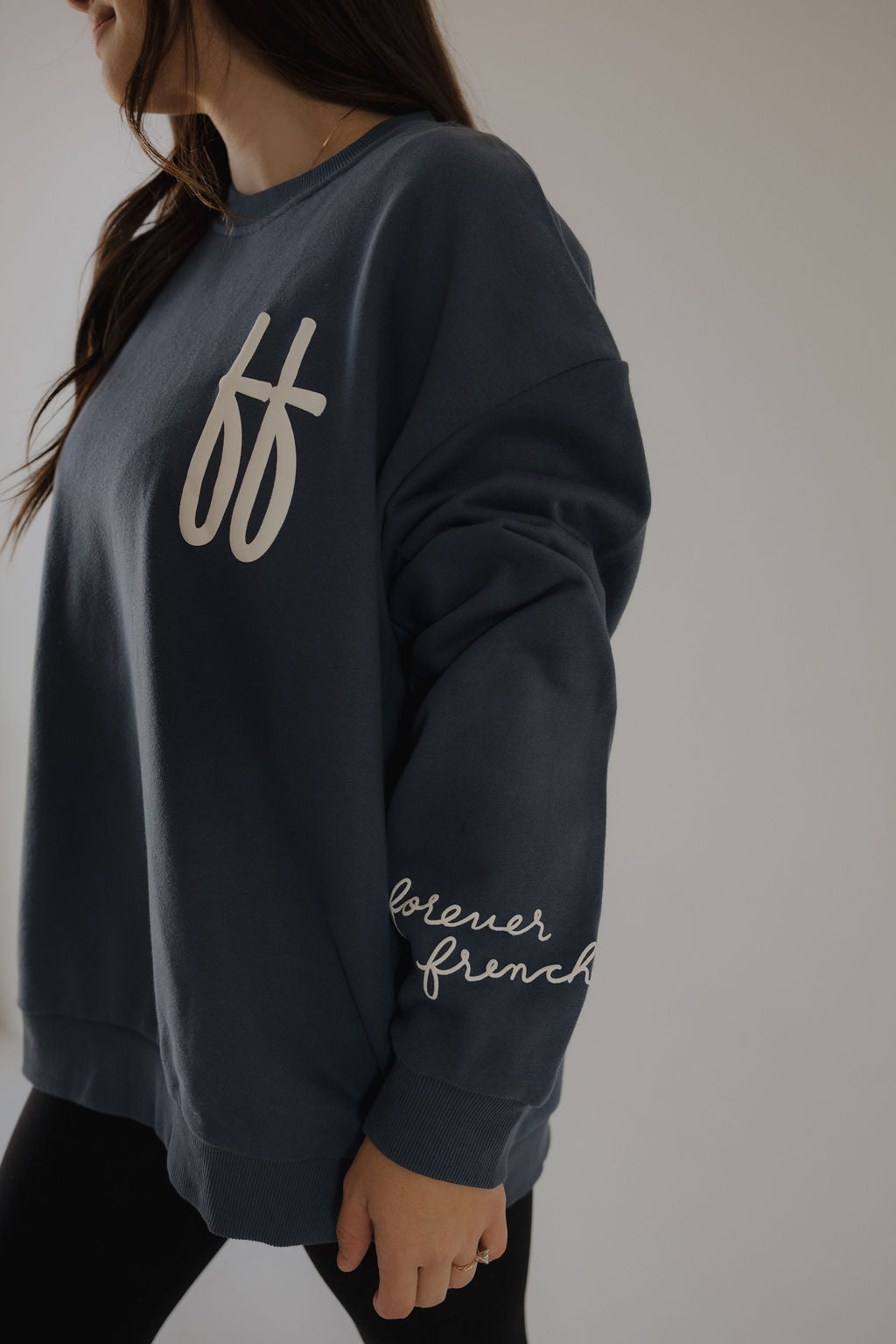 A woman wears the Adult Sweatshirt | ff Blue by forever french baby, featuring "ff" on the chest and "forever french" on the sleeve, standing against a plain background.