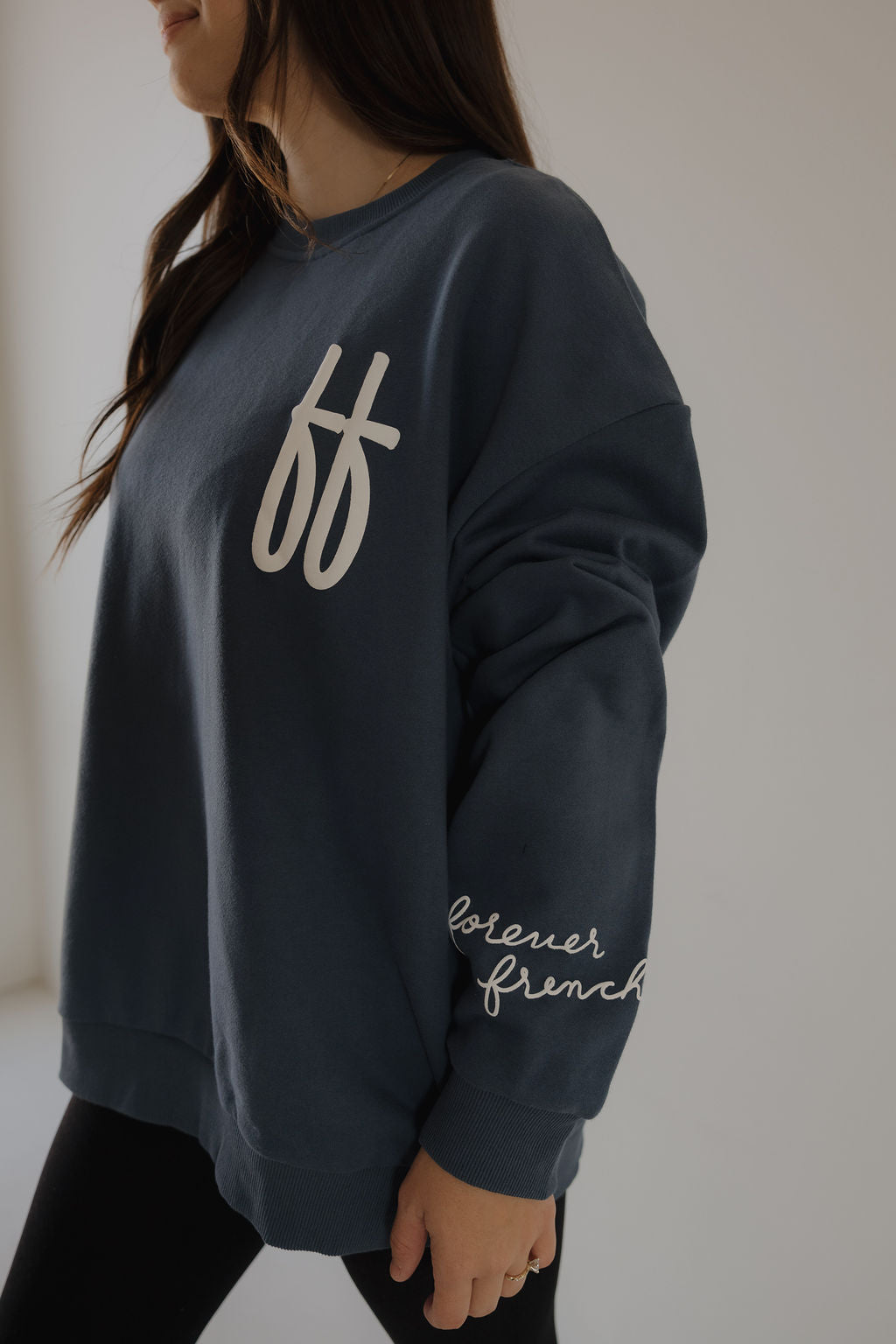 A woman wears the Adult Sweatshirt | ff Blue by forever french baby, featuring "ff" on the chest and "forever french" on the sleeve, in a relaxed fit. Only her lower face and upper body are seen in a softly lit room.