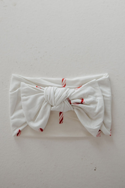 The Bamboo Head Wrap | Candy Cane by forever french baby is a white baby head wrap with red candy cane stripes and a large knotted bow, shown on a neutral background. A cute and popular accessory for little ones.