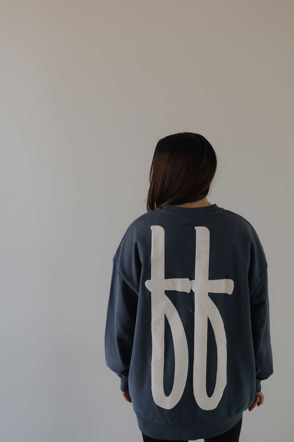 A person with long brown hair, seen from behind, wears the forever french baby Adult Sweatshirt | ff Blue with bold white abstract symbols on the back, standing against a plain light gray background.
