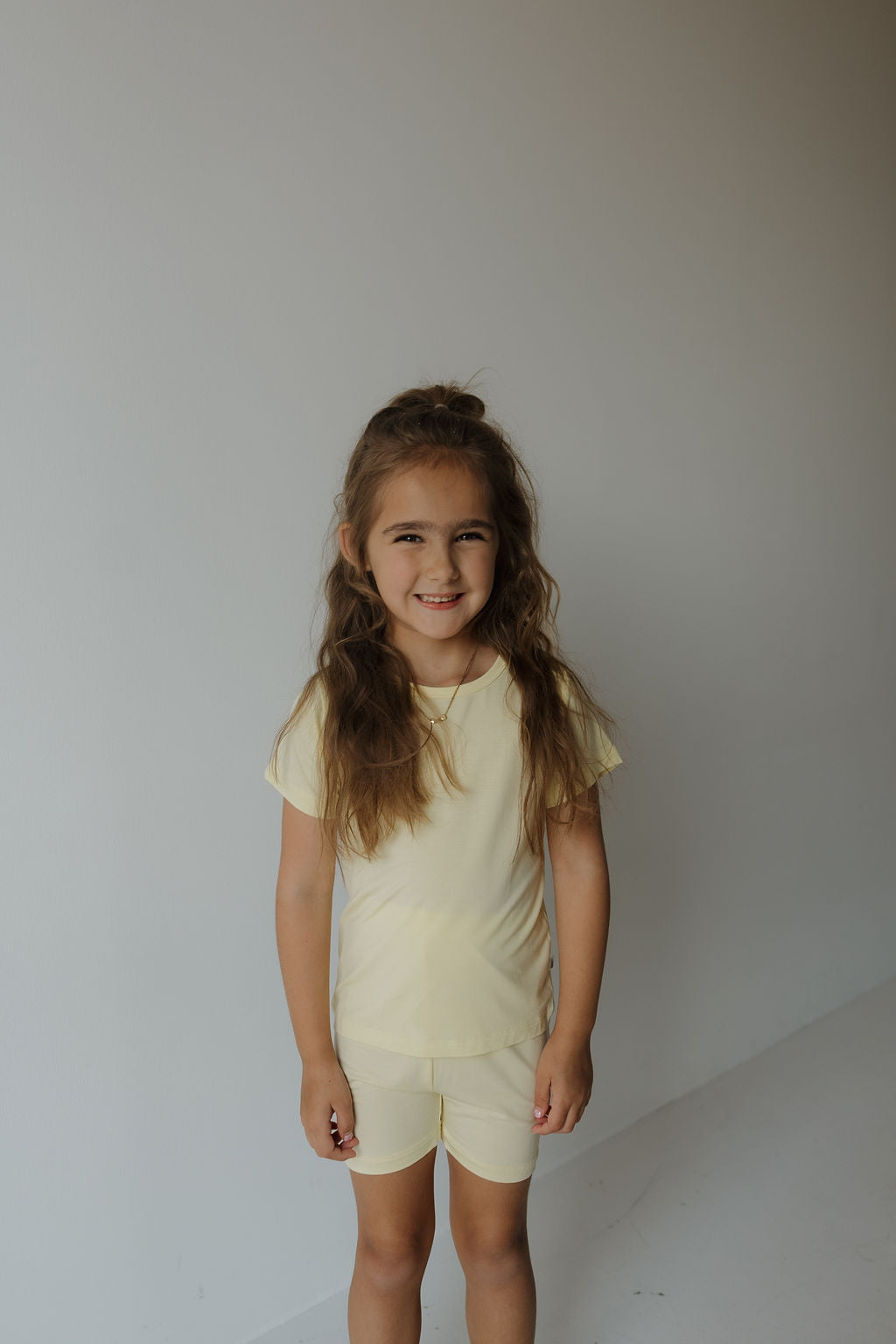 A young girl with long brown hair, wearing the forever french baby Bamboo Short Set | Sunny in light yellow, stands smiling against a plain, light-colored wall.