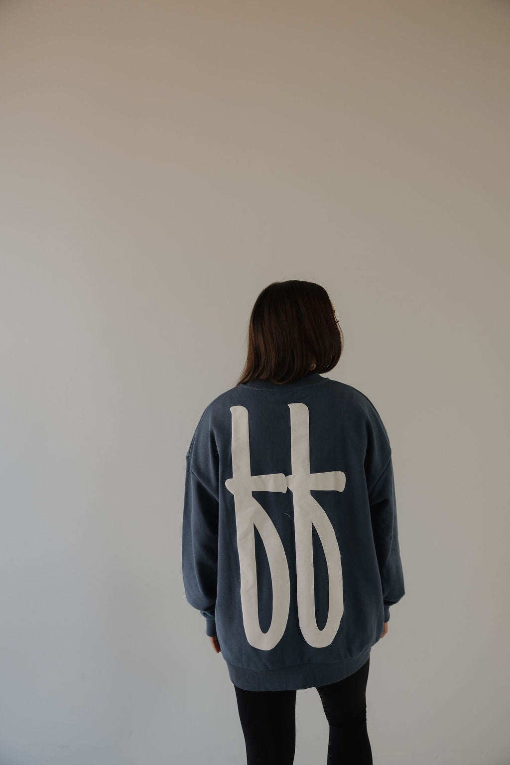 A person with straight dark hair faces a plain light wall, wearing the forever french baby Adult Sweatshirt | ff Blue, a relaxed fit in blue featuring large white graphic symbols on the back.