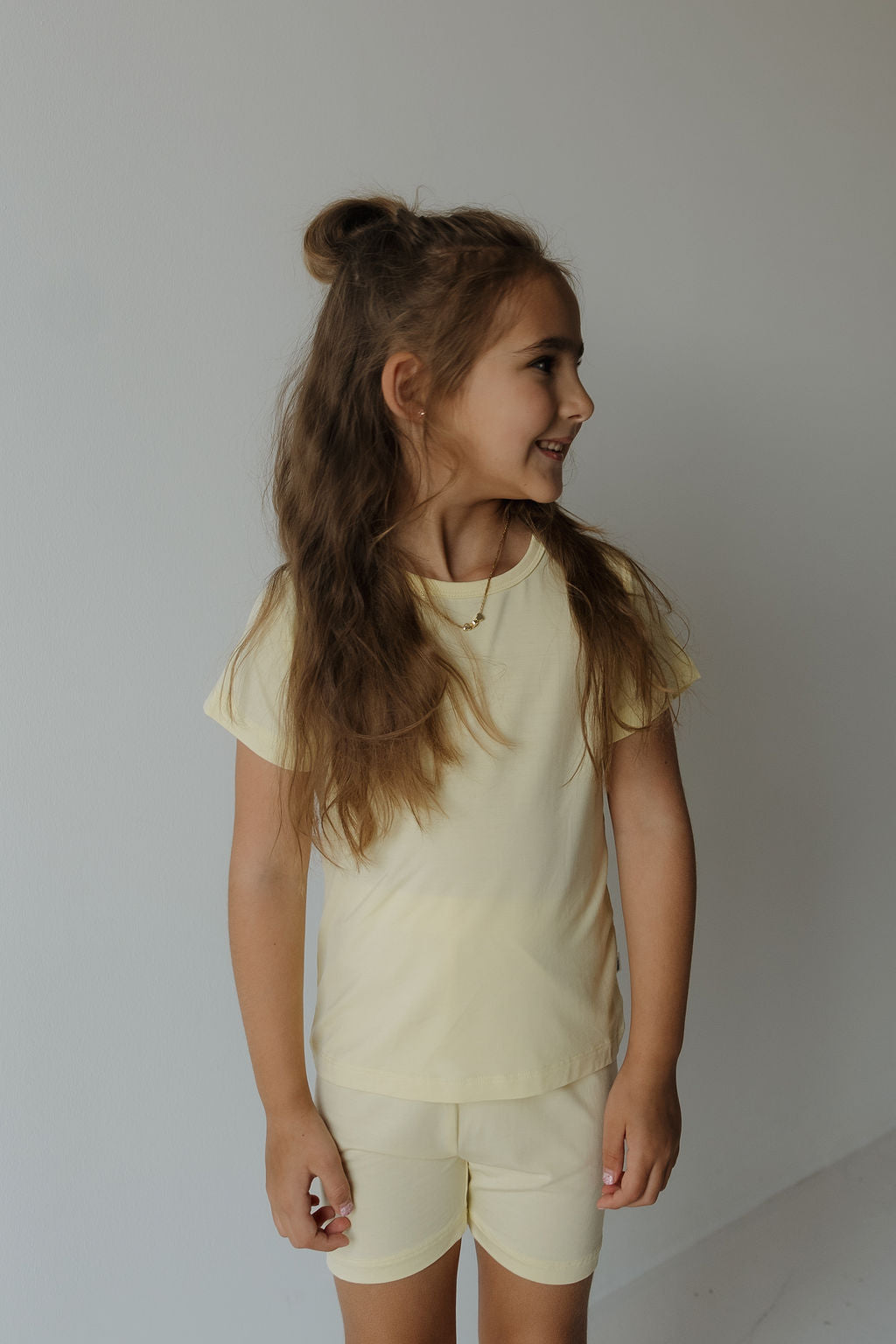 A young girl with long brown hair in a half-up style is wearing the forever french baby Bamboo Short Set | Sunny—a light yellow, breathable and comfy t-shirt with matching shorts—standing against a plain, light background and smiling as she looks to the side.