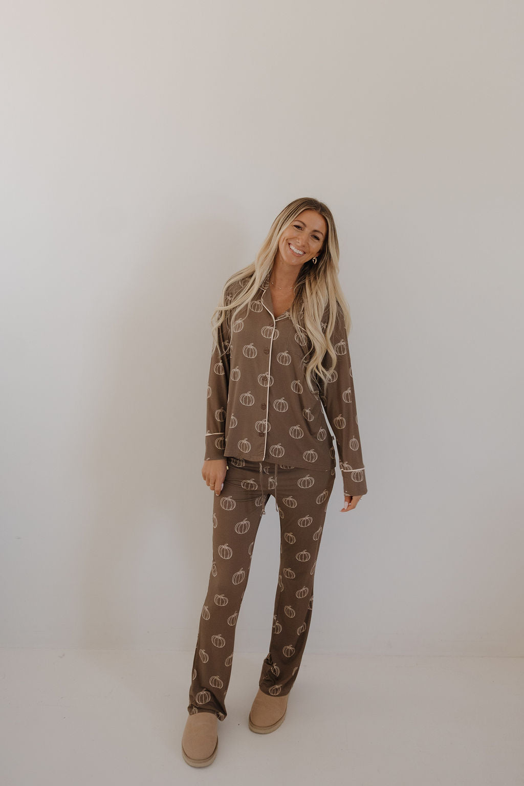 A woman with long blonde hair stands against a light wall, smiling and wearing the forever french baby Women's Bamboo Button Up Lounge Set in Brown Pumpkin, paired with beige slippers, highlighting its cozy comfort.