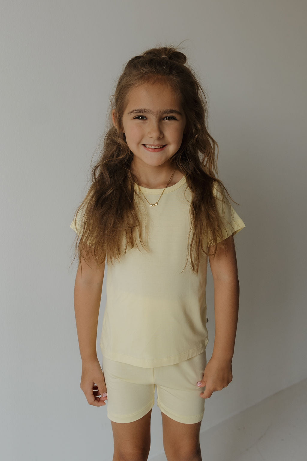 A young girl with long brown hair stands smiling against a plain light-colored wall, wearing the Forever French Baby Bamboo Short Set in Sunny.
