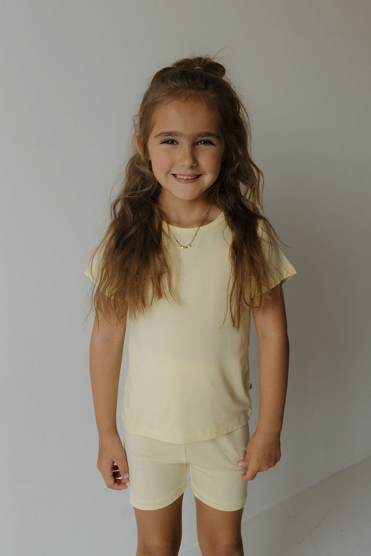 A young girl with long brown hair stands smiling against a plain light-colored wall, wearing the Forever French Baby Bamboo Short Set in Sunny.