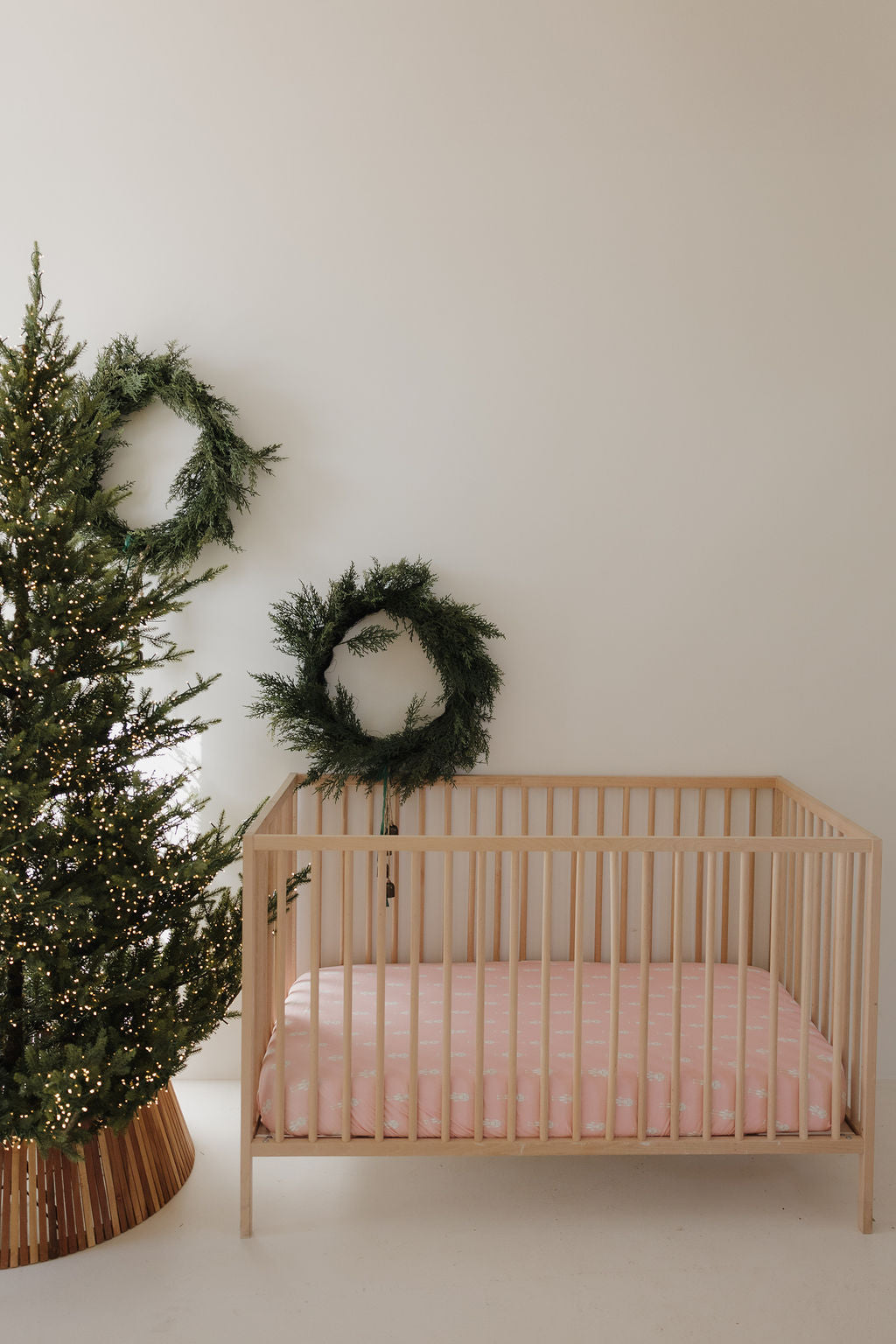 A minimalist nursery showcases a light wooden crib with the forever french baby Bamboo Crib Sheet | Nutcracker, a lit Christmas tree, and two green wreaths on a plain white wall.