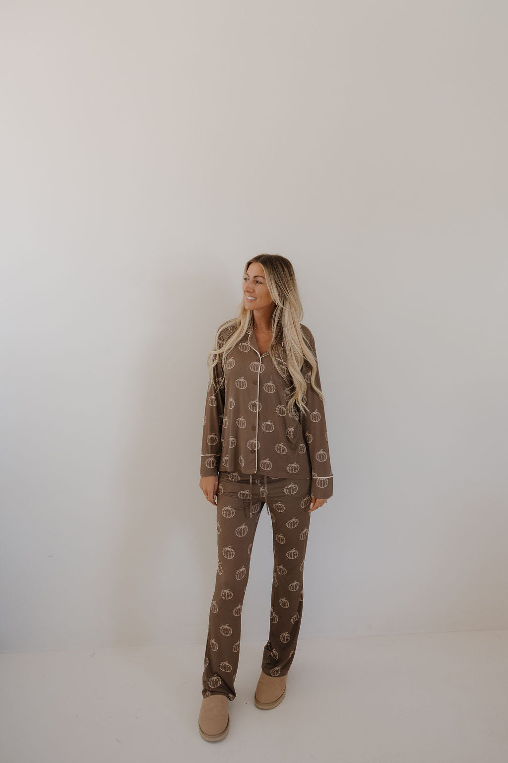 A woman with long blonde hair stands against a plain light background, smiling and looking to the side. She is wearing the forever french baby Women's Bamboo Button Up Lounge Set in Brown Pumpkin and tan slippers.