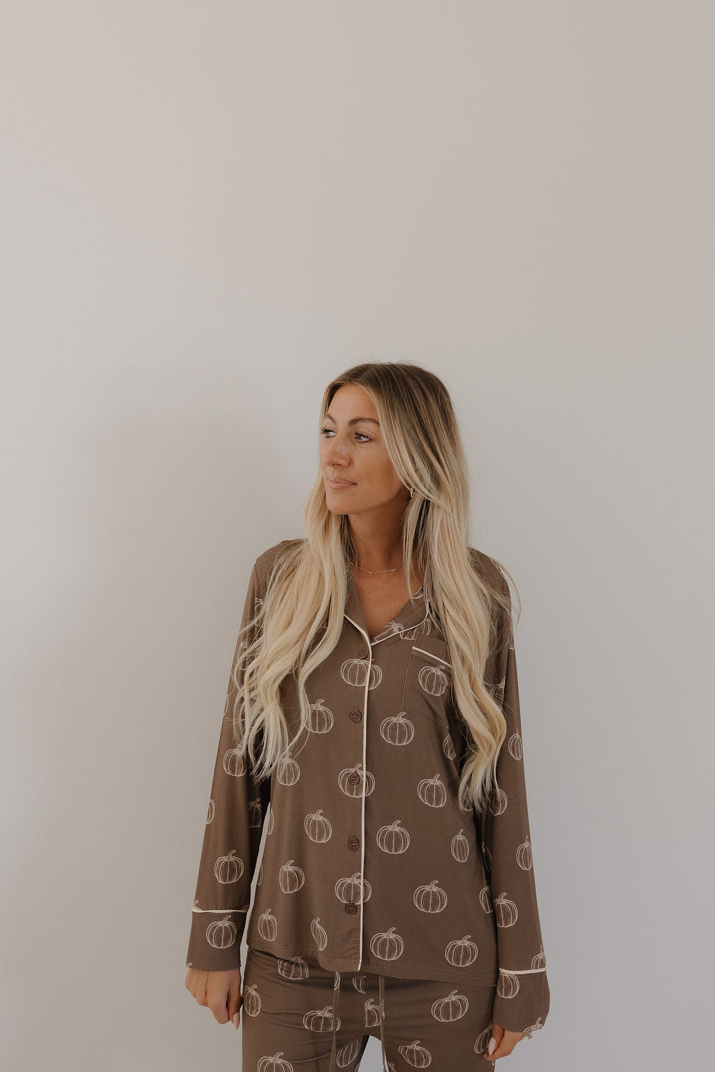 A woman with long blonde hair models the forever french baby Women's Bamboo Button Up Lounge Set in Brown Pumpkin, featuring a white pumpkin print, standing against a light background and gazing slightly left.