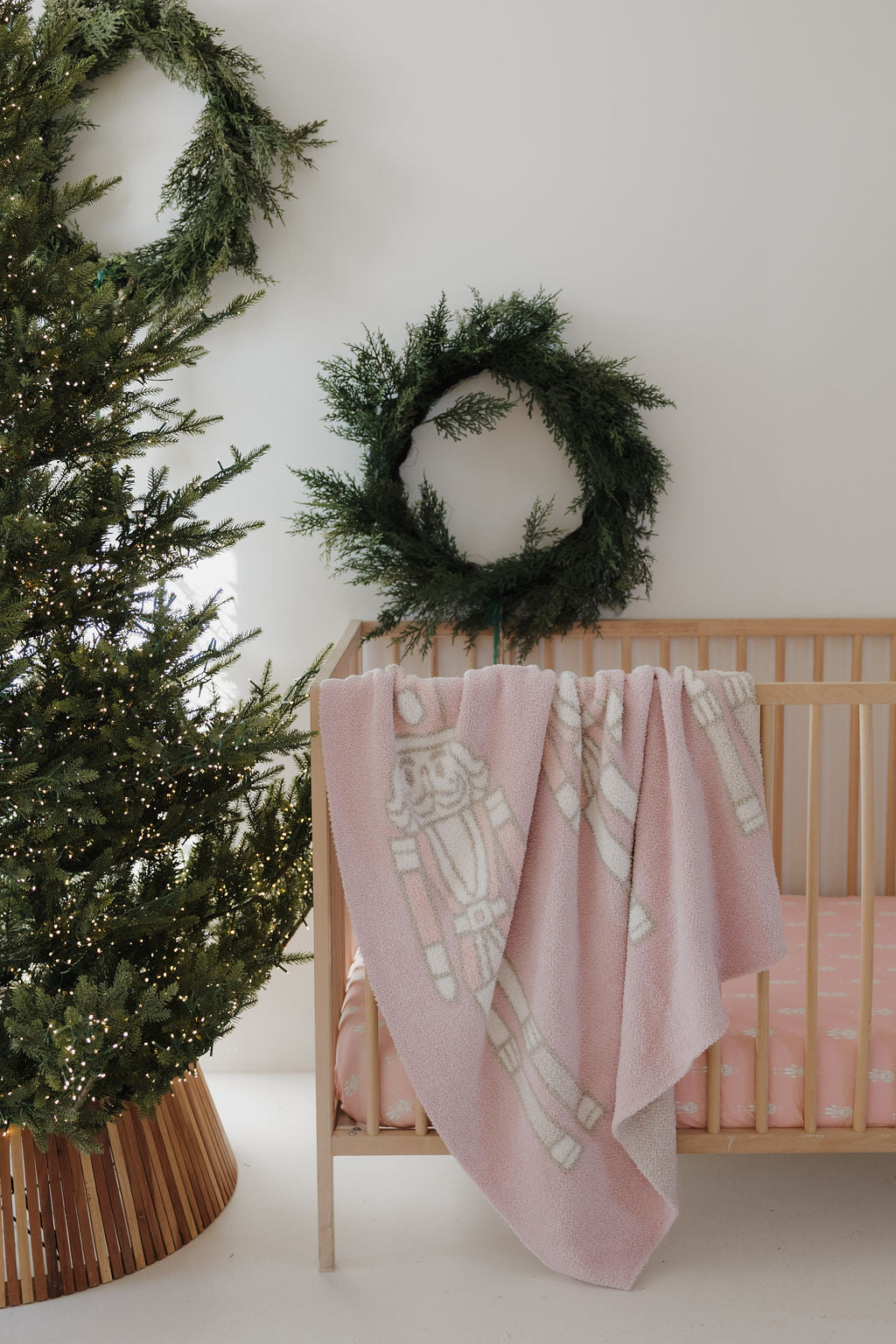 A cozy nursery with a wooden crib draped in the forever french baby Plush Blanket | Nutcracker, Oeko-Tex certified in pink with white nutcracker designs. Nearby, a Christmas tree and green wreath add to the festive atmosphere.