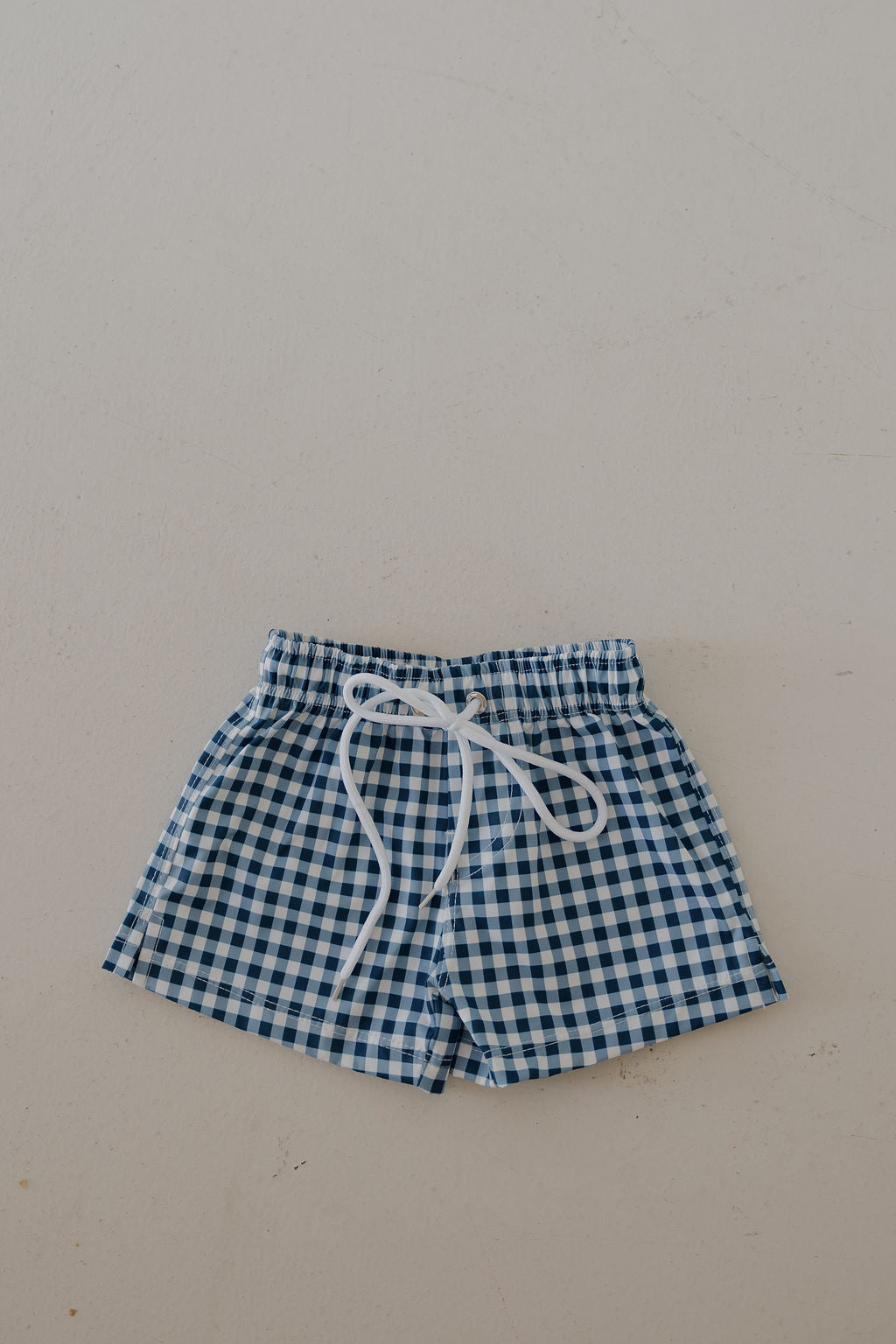 The forever french baby Child Board Short in blue gingham, featuring an elastic waistband and white drawstring, is perfect for summer and shown on a light, textured surface.