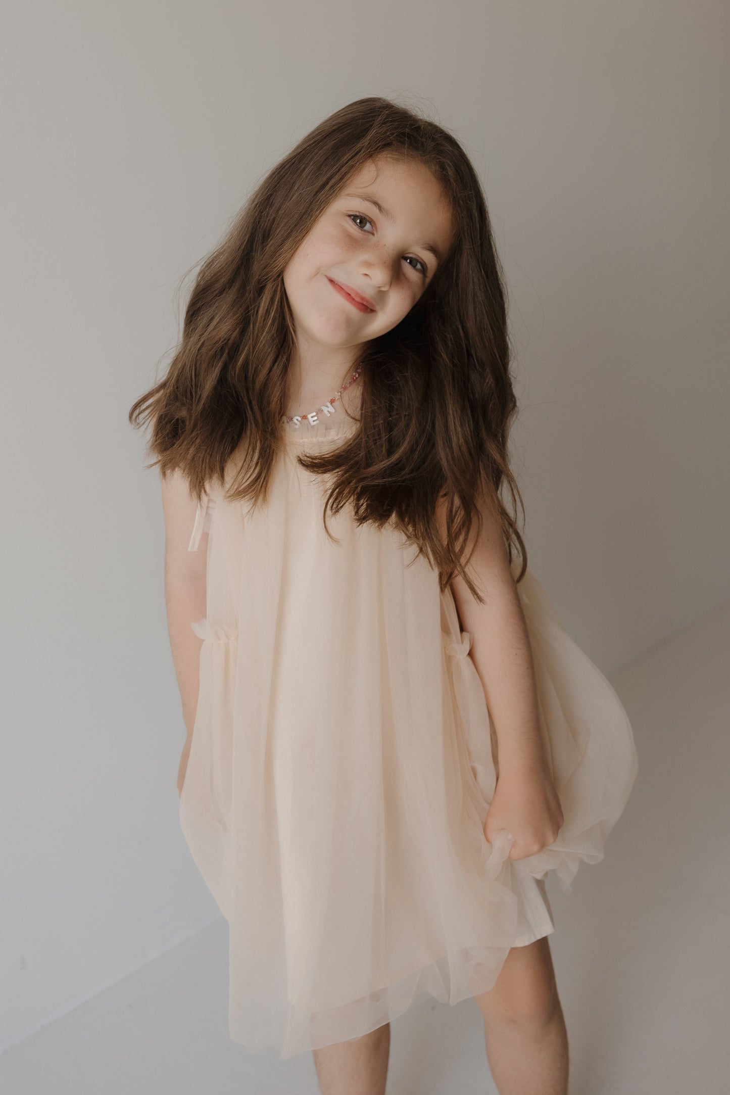 A young girl with long brown hair gently smiles as she stands against a plain, light background, wearing the forever french baby Tutu Dress | Slipper—a light, sleeveless design.