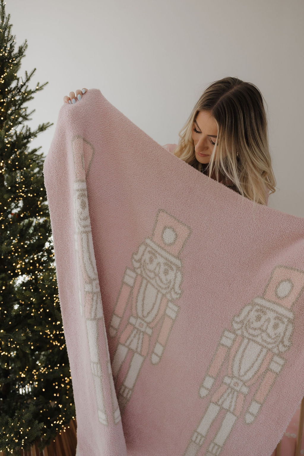 A woman holds up the forever french baby Plush Blanket | Nutcracker, a soft Oeko-Tex certified pink blanket with white and pink nutcracker designs. A decorated Christmas tree with string lights stands nearby against a plain white wall.