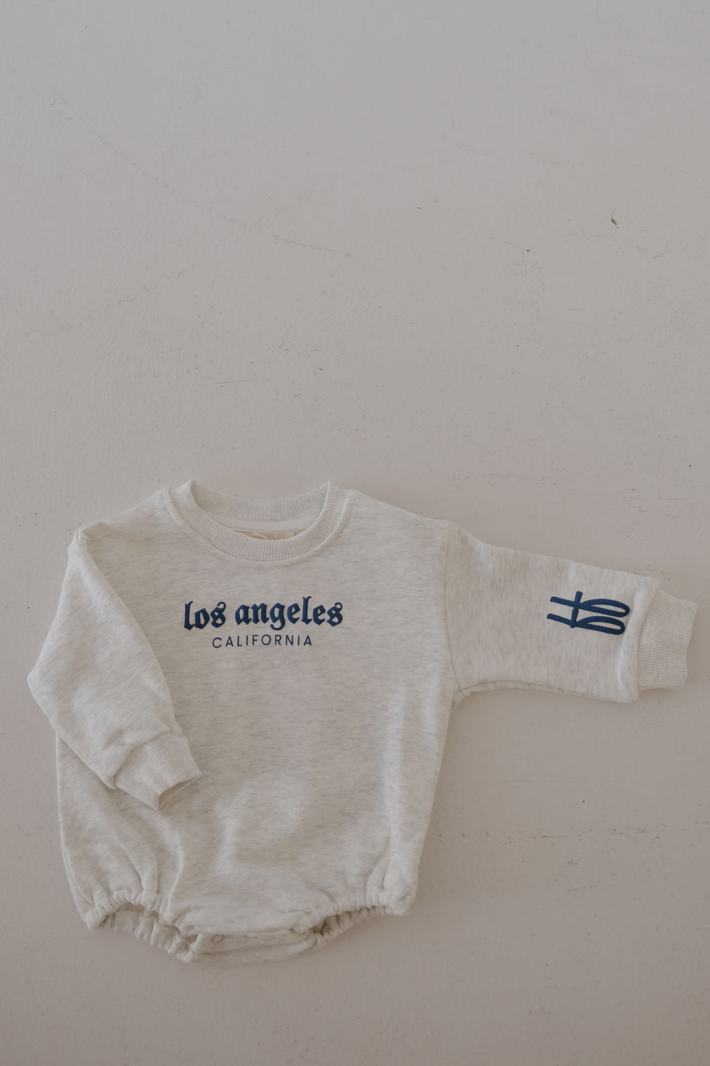The forever french baby Sweatshirt Romper | ff Los Angeles is a light gray romper with "Los Angeles California" in blue on the chest and a blue graphic on the left sleeve, shown laid flat on a neutral background.
