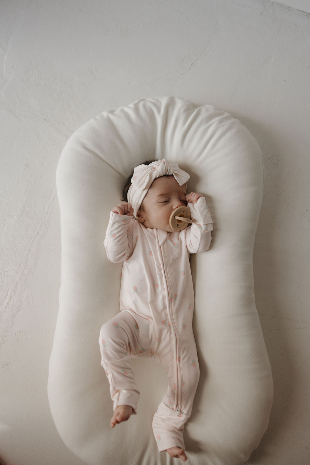 A baby in forever french baby’s Bamboo Zip Pajamas | Rosie—a pink bamboo sleepwear onesie—sleeps on a white cushioned lounger, holding a beige pacifier and wearing a matching headband. The background features a light, textured surface.