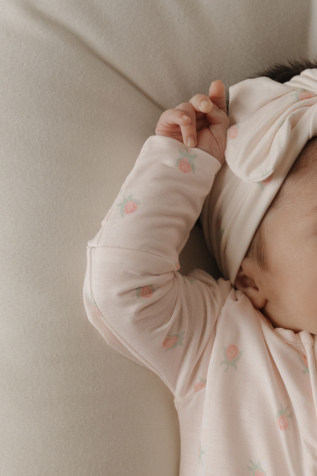 A baby wearing the forever french baby Bamboo Zip Pajamas | Rosie—a soft pink floral sleepwear set—along with a matching headband, lies on a cream-colored surface, one arm raised and only part of their face visible.