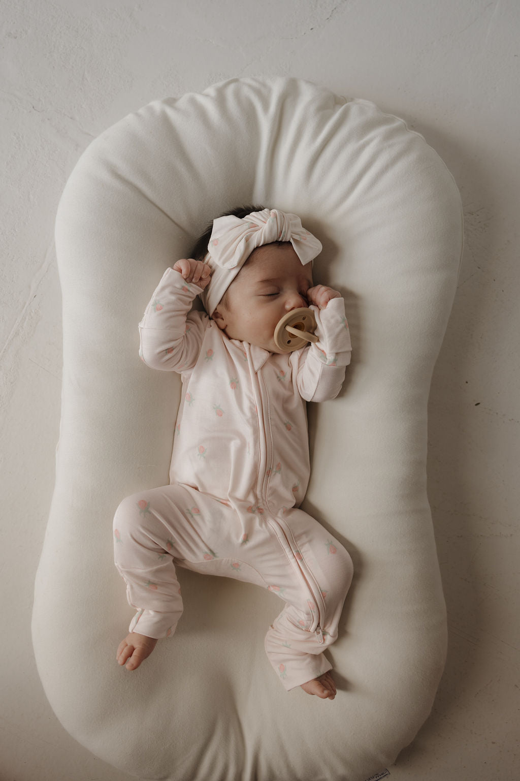 A baby wearing a light pink onesie and the forever french baby Bamboo Head Wrap | Rosie, featuring an adjustable bow, sleeps peacefully on a soft white cushion while holding a pacifier.